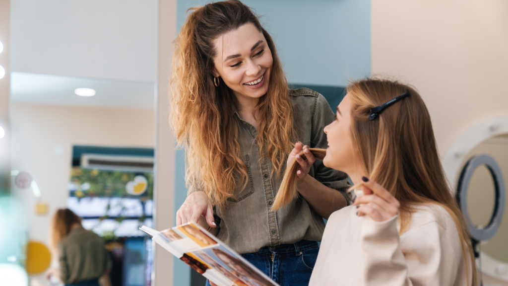 Hairstylist and client discussing hairstyle options while reviewing a magazine, both smiling and engaged in a conversation about the perfect hairstyle.